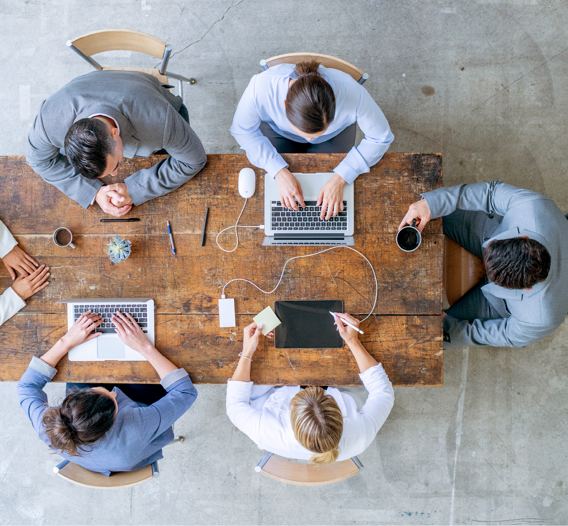 An overhead view of a group of businessmen and businesswomen at a conference table for a meeting together. They are using electronics such as a laptop.